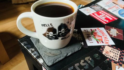 Eine Kaffeetasse mit Totenkopf- und Glockenmotiven und der Aufschrift „HELLS BELLS ST. PAULI“ steht auf einem Schieferuntersetzer. Die Tasse steht auf einem Tisch, der mit verschiedenen Aufklebern bedeckt ist, darunter auch einige mit Bezug zu St. Pauli.