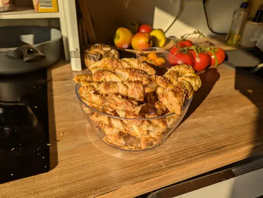 photo of a transparent plastic bowl filled with many twisted onion sticks. they have a diameter of about 3 to 4 centimeters (about 1.5 inches) and they have bits of cheese on them. they appear golden brown and like they came right out of the oven (whivh they did). in the background there are some tomatos, apples and a pan over an induction stove. the lighting is very warm, with sunlight lighting the whole kitchen scenery.