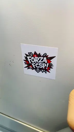 Ein Aufkleber mit dem Schriftzug „FORZA FCSP!“ in fetter, stilisierter Schriftart, umgeben von roten und schwarzen Grafikelementen, darunter Ketten und Totenköpfe. Der Aufkleber klebt auf einer hellen Wand.