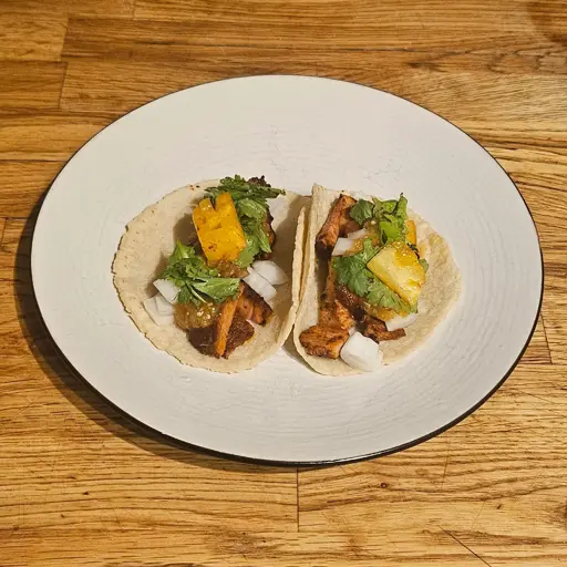 A plate of two tortillas filled with fried marinated pork, chopped white onion, salsa verde, cilantro and pineapple
