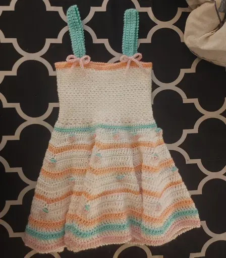 Crochet children's dress. White with blue-green, orange, and pink stripes and four rows of strawberries between the orange stripes. Blue-green straps with pink bows where the straps attach to the dress.