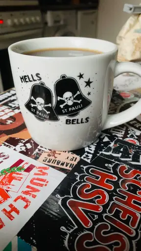 Eine Kaffeetasse mit einem Motiv aus zwei Glocken und Totenköpfen, begleitet von den Aufschriften „HELLS BELLS“ und „ST. PAULI“. Die Tasse steht auf einem Tisch, der mit verschiedenen Aufklebern und Verzierungen bedeckt ist.