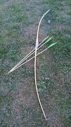 Auf der Wiese vor uns liegen ein fertiger Holzbogen und drei Holzpfeile, die eine grün und orangende Befiederung haben Auf der Wiese vor uns liegen ein fertiger Holzbogen und drei Holzpfeile, die eine grün und orangende Befiederung haben