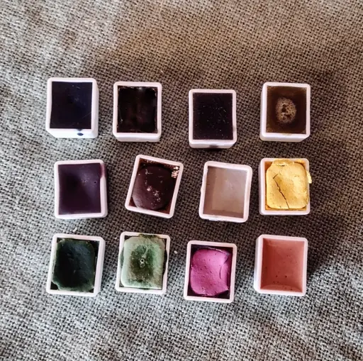 An arrangement of twelve watercolour pots filled with different coloured paints on a green tablecloth.