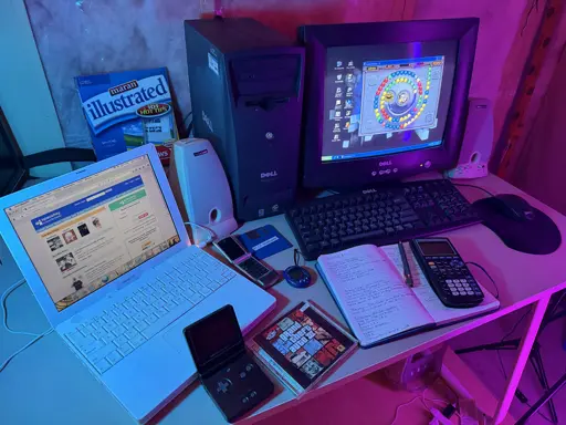 A staged early-2000s computer desk setup lit with blue and magenta accent lighting. On the left, a white iBook G4 laptop displays SpaceHey, a modern MySpace-style social network, in a Firefox-based browser. In the center is a Dell Dimension 2200 tower connected to a Dell CRT monitor showing Windows XP with the game Zuma Deluxe running, using a Dell keyboard and mouse. Beige Harman/Kardon desktop speakers sit beside the monitor. Spread across the desk are period accessories including a Motorola Razr flip phone, a blue Tamagotchi Connection, a TI-84 graphing calculator resting on an open handwritten notepad, a black Game Boy Advance SP, and a blue 3.5-inch floppy disk labeled “Plop Bootloader.” A GTA III jewel case lies near the front edge of the desk, and a Maran Illustrated Windows XP: 101 Tips book stands behind the computers.