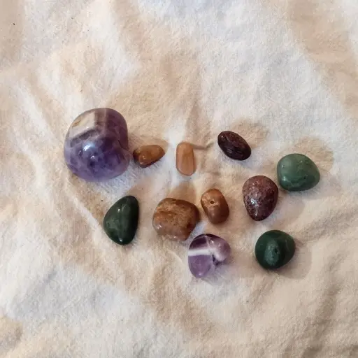 A hundful of different, smoothed rocks and minerals including amethyst, citine, jade and unidentified pieces, one a white cloth.
